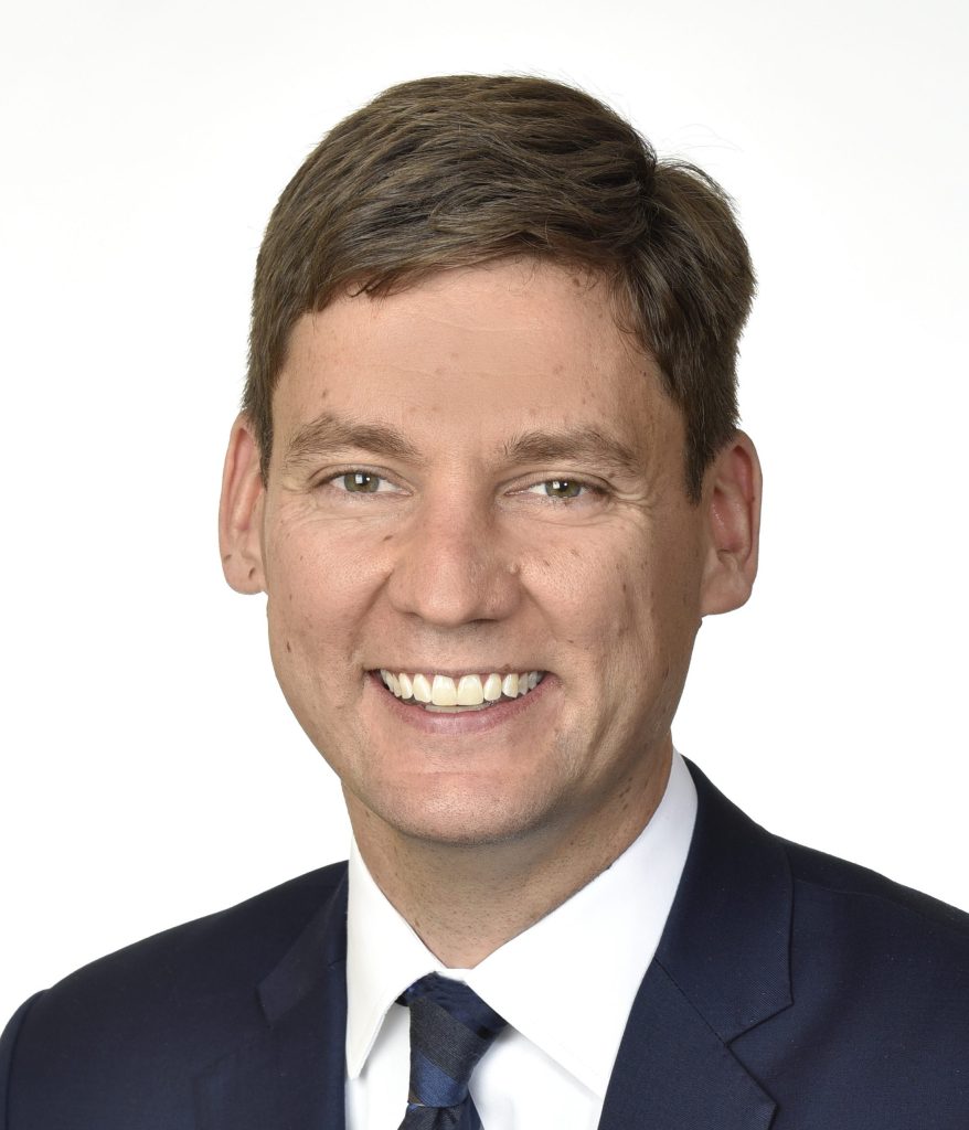 A man in a dark suit with a white shirt and tie smiles against a plain white background. His hair is short and brown.