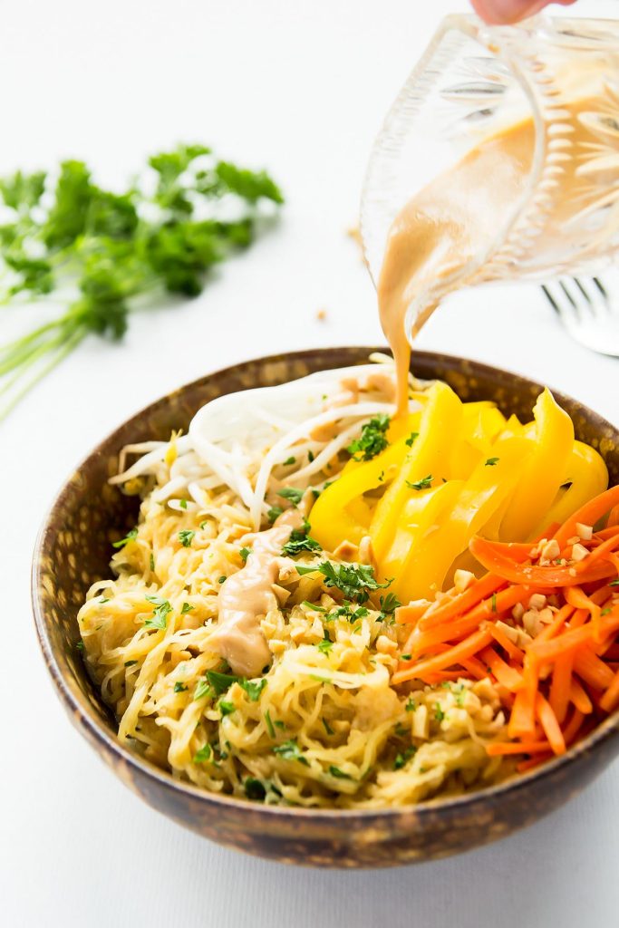 A bowl of noodles topped with sliced yellow bell peppers, carrots, bean sprouts, and herbs. Peanut sauce is being poured over the dish. A bunch of parsley and a fork rest blurred in the background.