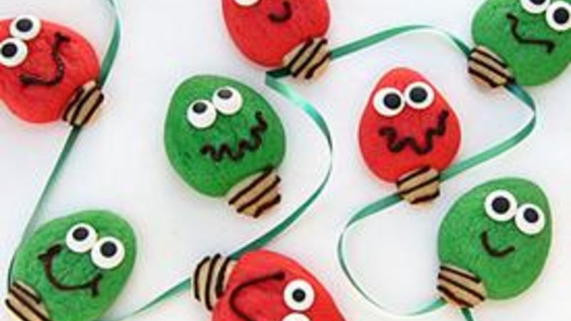 Colorful cookies shaped like Christmas lights in red and green with cartoonish faces, googly eyes, and frosting accents, arranged with a green ribbon on a white surface.