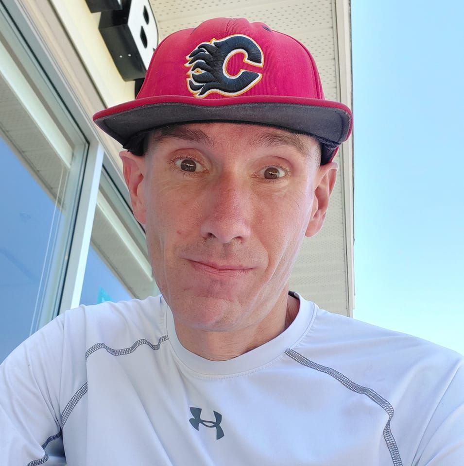 A person in a red baseball cap with a flaming C logo and a white Under Armour shirt takes a selfie outdoors. The sky is clear, and part of a building is visible in the background.