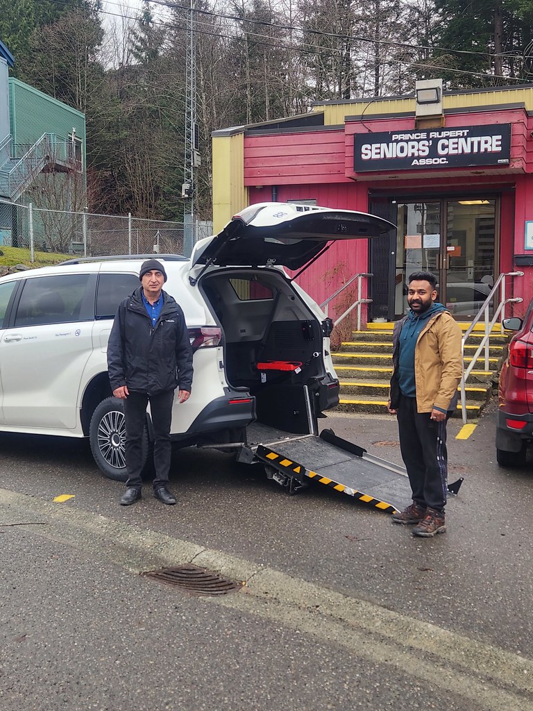 Two men stand beside a white vehicle with an open ramp leading to its back, parked in front of a building labeled Prince Rupert Seniors Centre. Trees and railings are visible in the background.