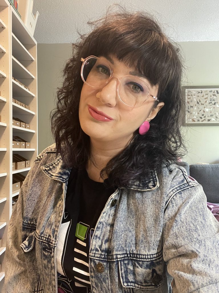 A person with wavy dark hair, wearing pink glasses and earrings, smiles slightly while wearing a denim jacket over a printed T-shirt. As a self-advocate in the DiversAbility space, they exude confidence indoors with shelves nearby and a framed picture on the wall behind.