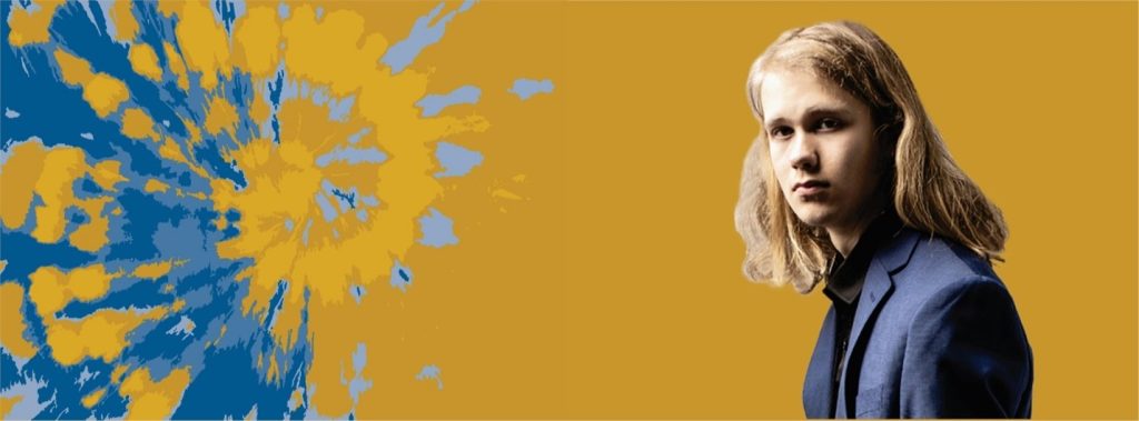 A person with long blond hair wearing a dark jacket stands against a mustard yellow background. To the left, a dynamic splash of blue, white, and yellow shapes forms an abstract pattern, embodying the spirit of an advocate for DiversAbility.