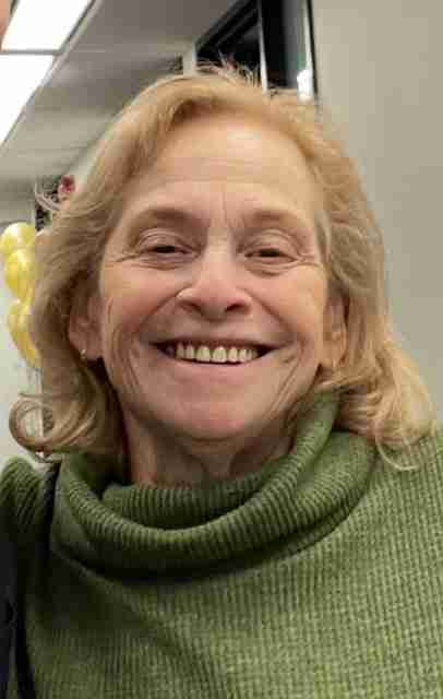 A smiling woman with light-colored hair and wearing a green turtleneck sweater stands indoors. Balloons are visible in the blurred background.