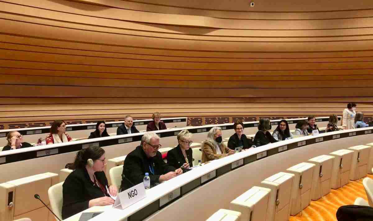 People sit in a curved conference room with wooden paneling, identified by nameplates. They are engaged in discussion, some using headsets. The setting suggests a formal meeting or event.