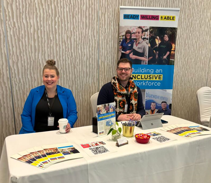 Two people sit at a booth with a Ready, Willing & Able banner. The woman wears a blue blazer and holds a coffee cup, while the man wears a colorful scarf. The table displays brochures, QR codes, and a bowl of candy.
