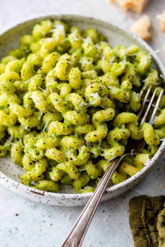A bowl of pasta covered in a creamy green pesto sauce, topped with grated cheese and cracked black pepper, with a fork resting on the side of the bowl.